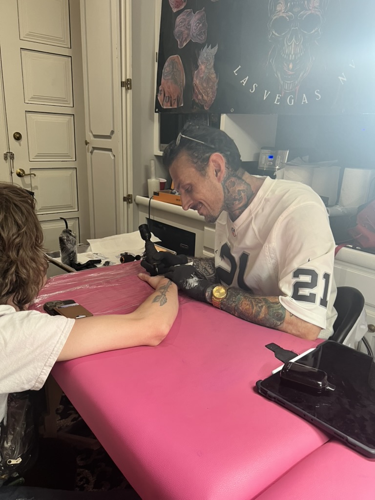 Joshua Cole tattoos black arrow on inner forearm at pink table with Vegas banner behind — Joshua Cole offsite booking, Work of Art Las Vegas