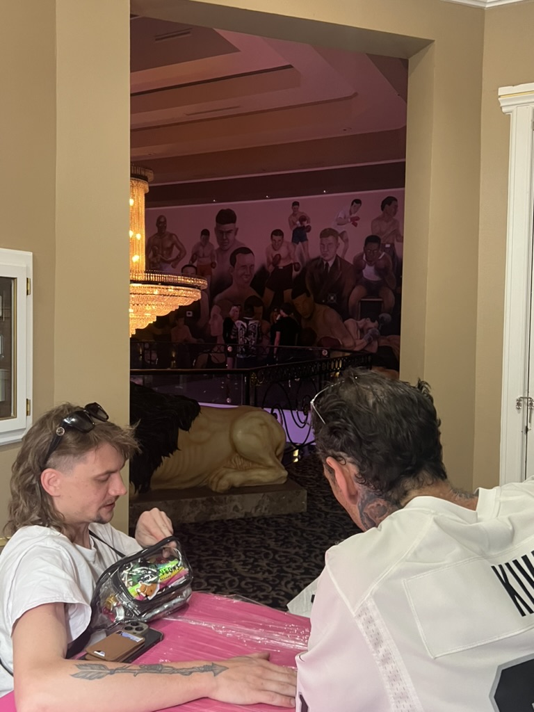 Men sit at pink offsite table; client shows fresh arrow forearm tattoo near lion statue — Joshua Cole offsite booking, Work of Art Las Vegas