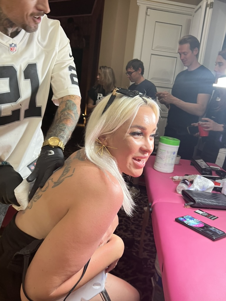Joshua Cole wipes fresh back tattoo on smiling blonde woman at crowded offsite setup — Joshua Cole offsite booking, Work of Art Las Vegas