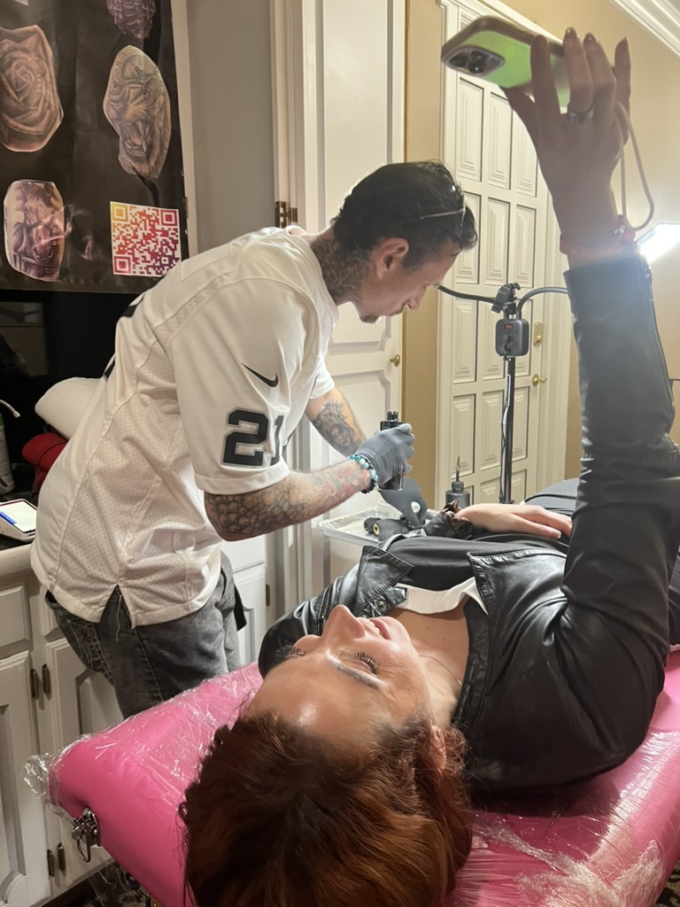 Client on pink table films Joshua Cole tattooing her arm with phone selfie — Joshua Cole offsite booking, Work of Art Las Vegas