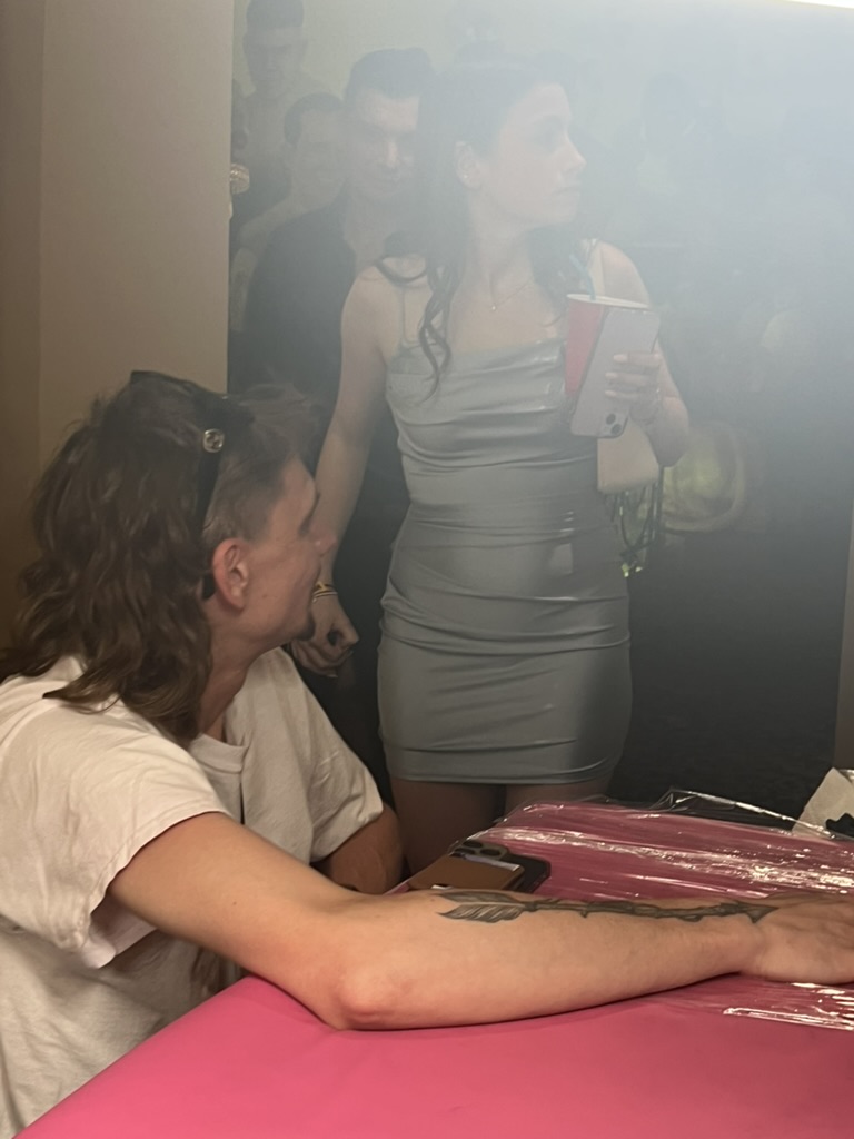 Client displays finished black arrow forearm tattoo at pink-covered offsite event table — Joshua Cole offsite booking, Work of Art Las Vegas