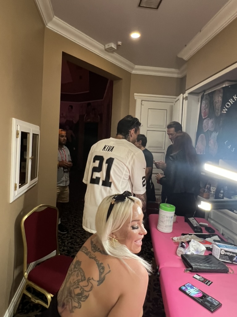 Blonde client turns showing finished ornate back tattoo at pink offsite event table — Joshua Cole offsite booking, Work of Art Las Vegas