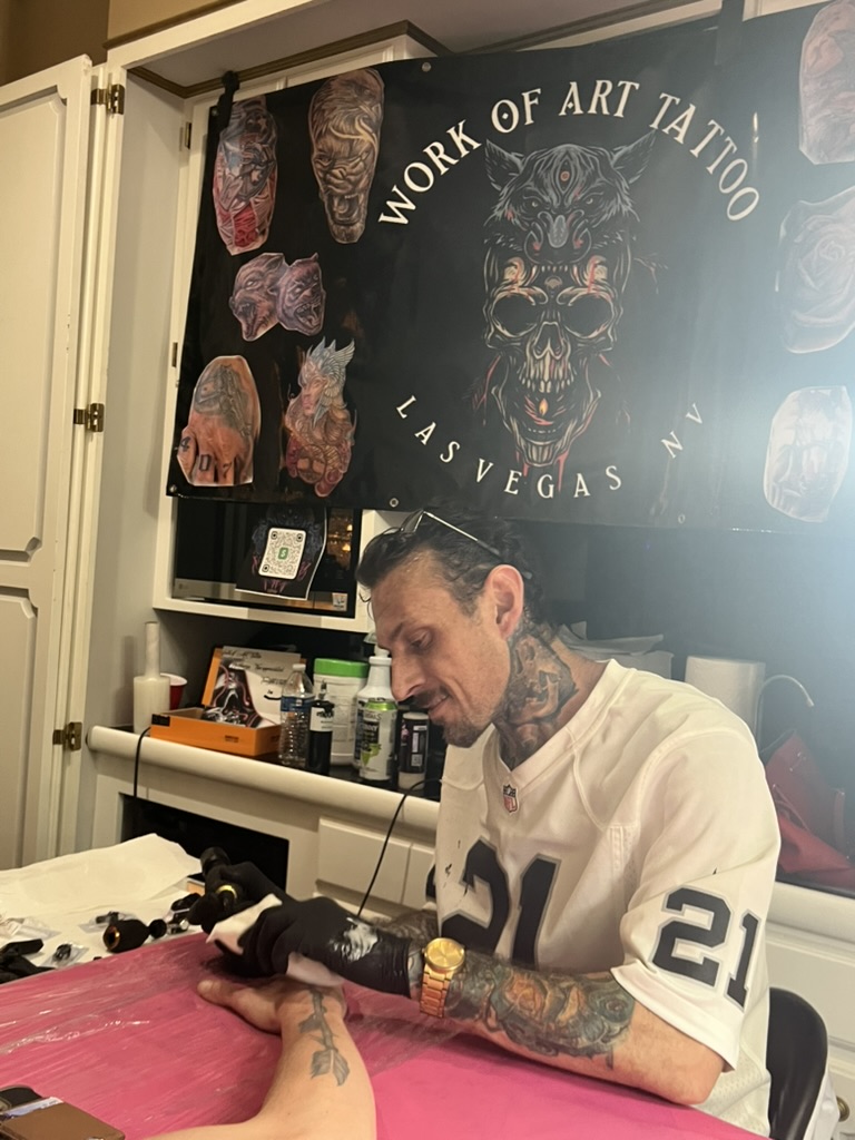 Joshua Cole tattoos forearm at pink table beneath Work of Art Las Vegas skull banner — Joshua Cole offsite booking, Work of Art Las Vegas