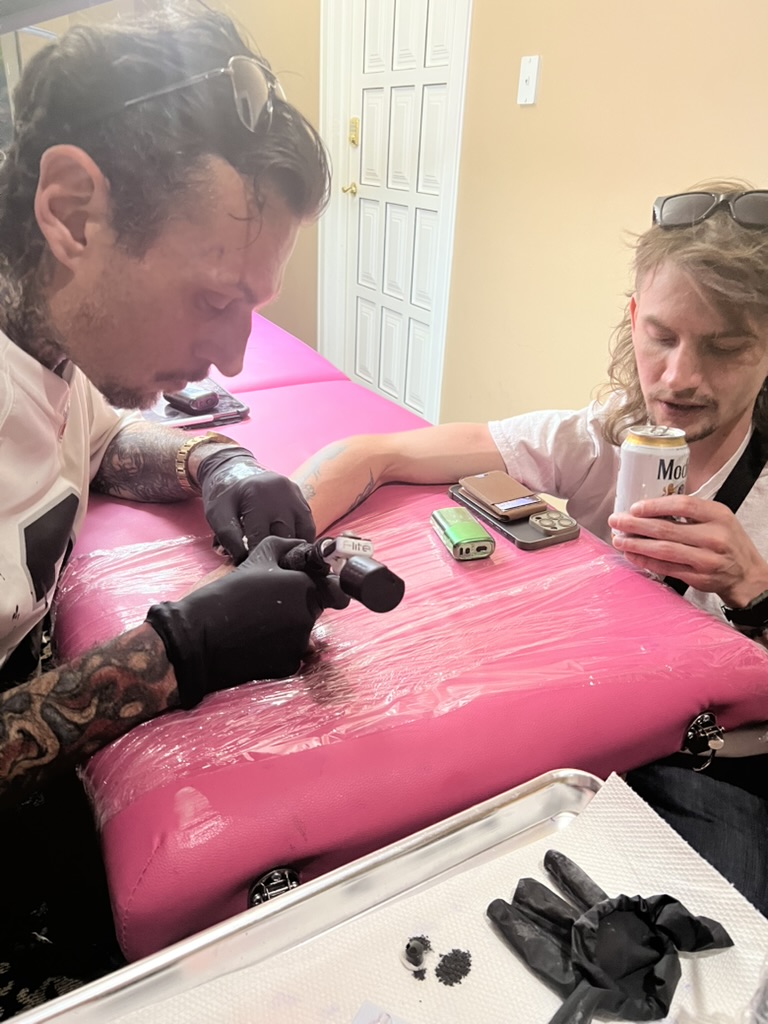 Joshua Cole tattoos forearm as mullet client holds Modelo on pink table — Joshua Cole offsite booking, Work of Art Las Vegas
