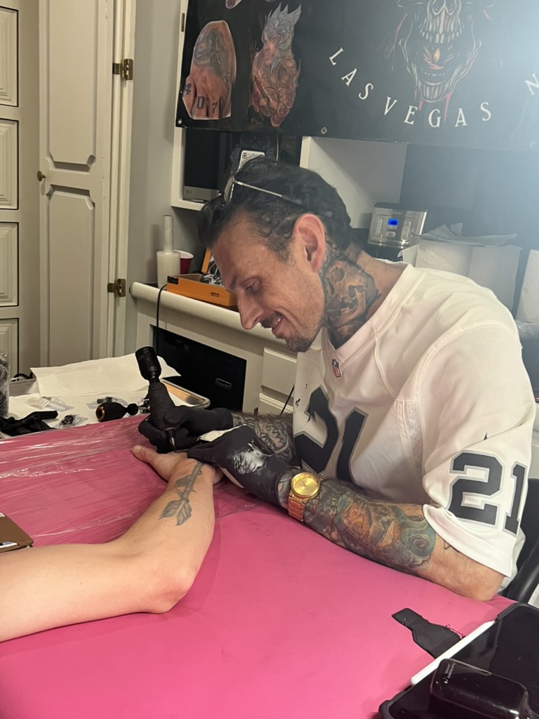 Joshua Cole tattoos back of hand on pink table; client has arrow forearm tattoo — Joshua Cole offsite booking, Work of Art Las Vegas