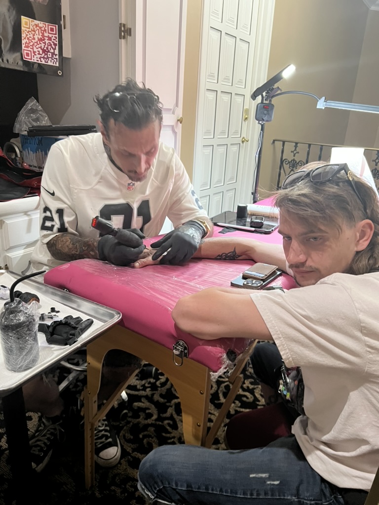 Joshua Cole tattoos client's hand on pink table; mullet client faces camera — Joshua Cole offsite booking, Work of Art Las Vegas