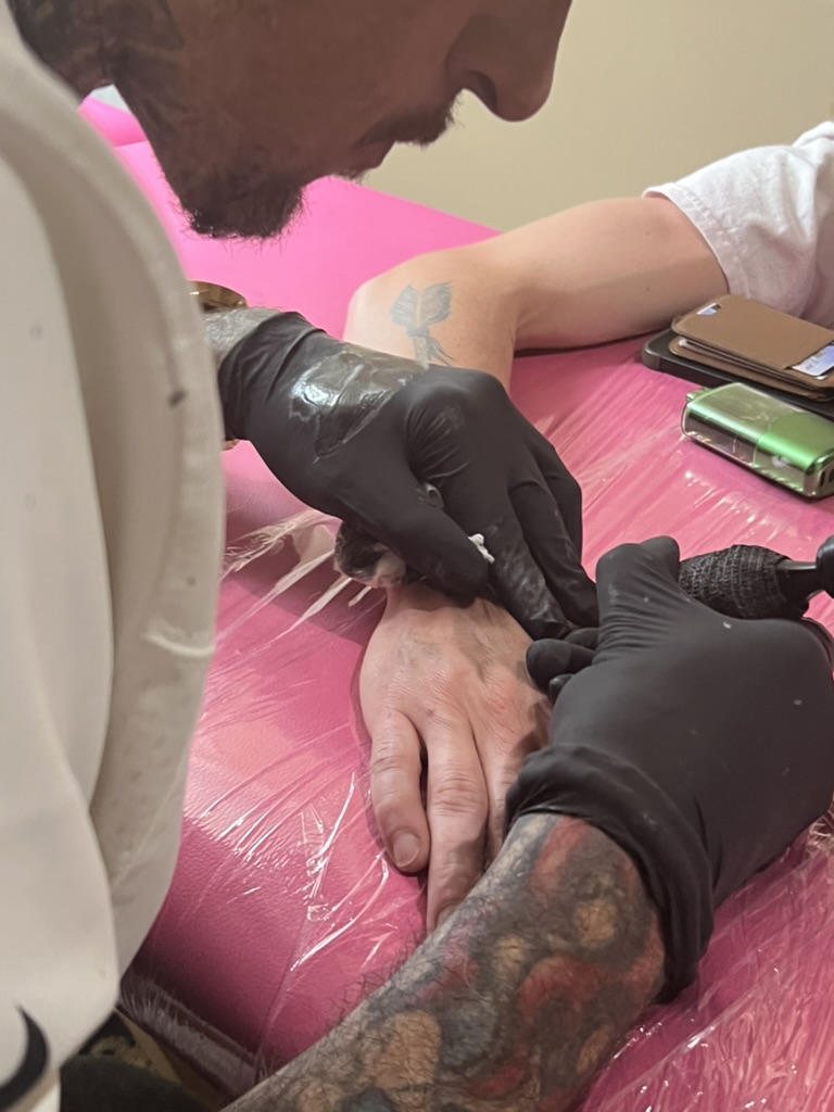Joshua Cole tattoos back of client's hand on pink-wrapped table; arrow on forearm — Joshua Cole offsite booking, Work of Art Las Vegas