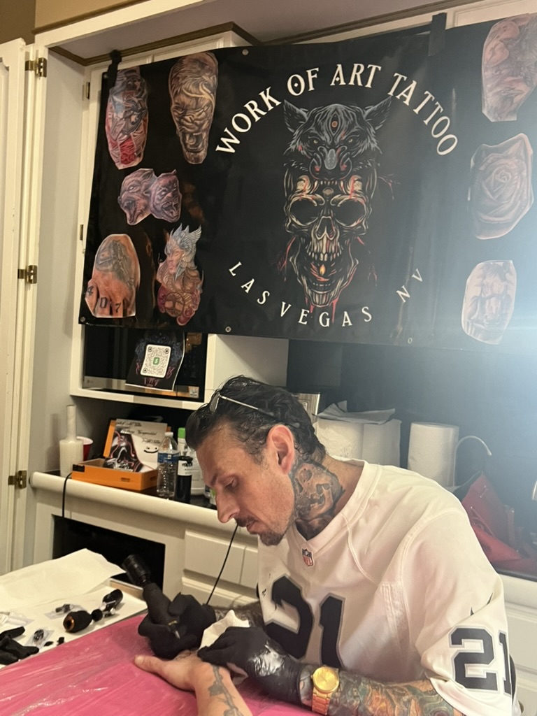 Joshua Cole tattoos client's hand on pink table beneath Work of Art Las Vegas banner — Joshua Cole offsite booking, Work of Art Las Vegas
