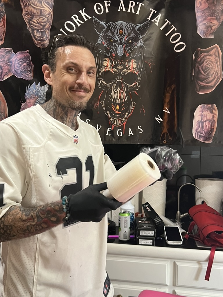 Joshua Cole grins holding clear barrier film roll at mobile banner — Joshua Cole offsite booking, Work of Art Las Vegas