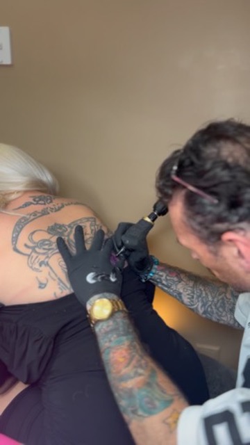 Joshua Cole inks ornate black-grey back tattoo on blonde client leaning forward — Joshua Cole offsite booking, Work of Art Las Vegas