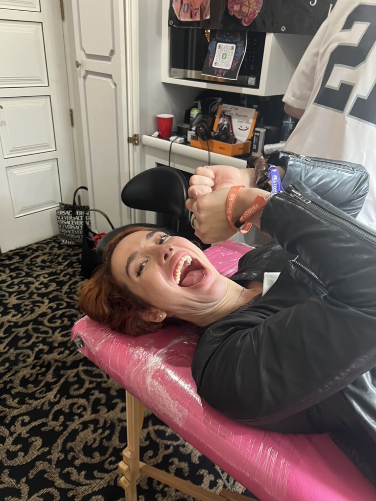Client grins wide on pink tattoo table with festival wristbands at VIP party — Joshua Cole offsite booking, Work of Art Las Vegas