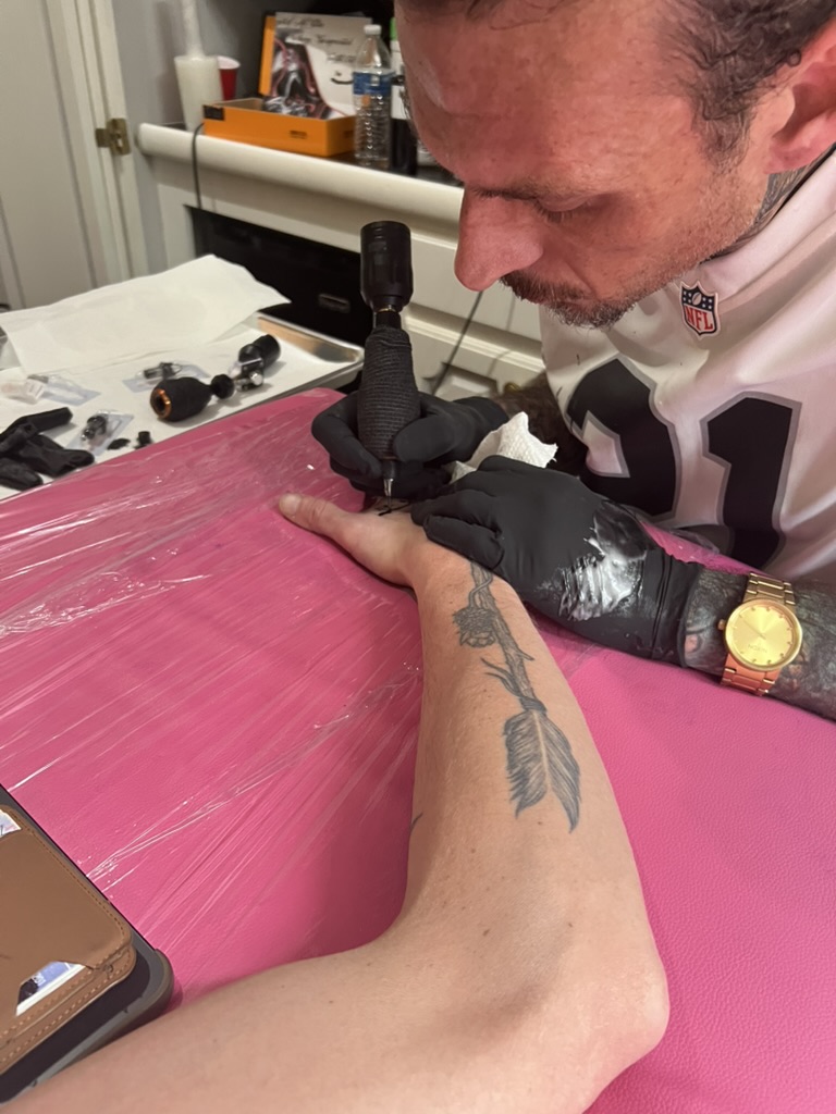 Joshua Cole tattoos small design on hand beside arrow-and-rose forearm at pink table — Joshua Cole offsite booking, Work of Art Las Vegas