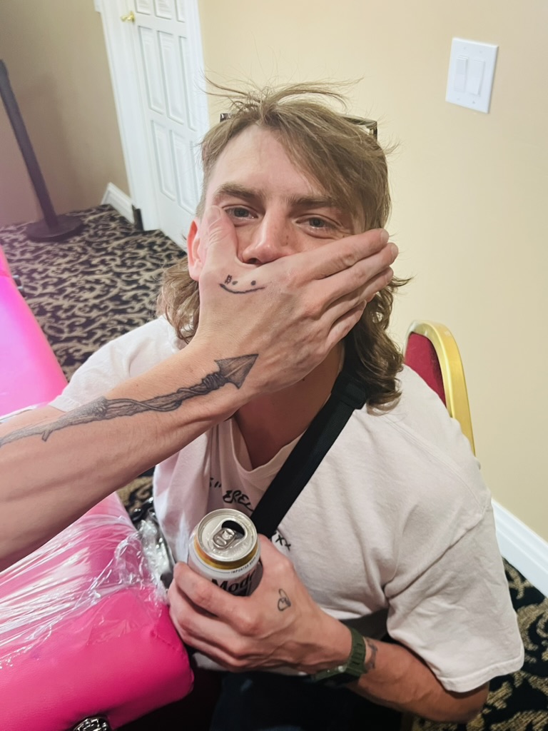 Smiley hand tattoo covers man's mouth beside spear forearm tattoo at pink offsite table — Joshua Cole offsite booking, Work of Art Las Vegas