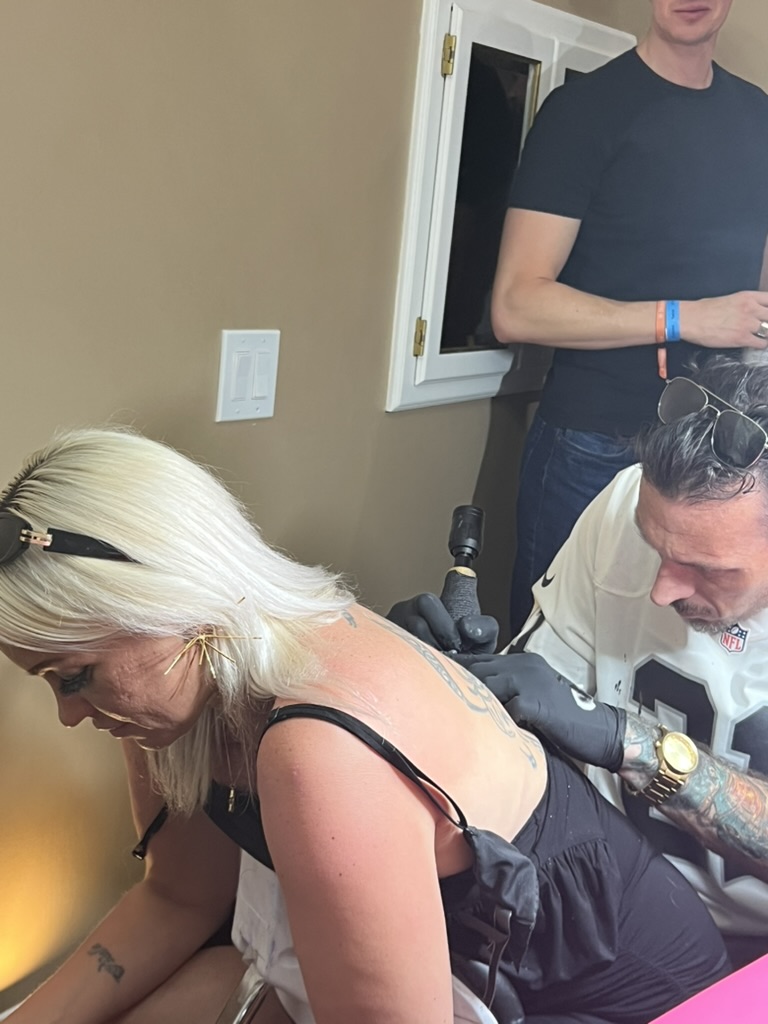 Joshua Cole tattoos upper-back piece on blonde client in Raiders jersey setup — Joshua Cole offsite booking, Work of Art Las Vegas