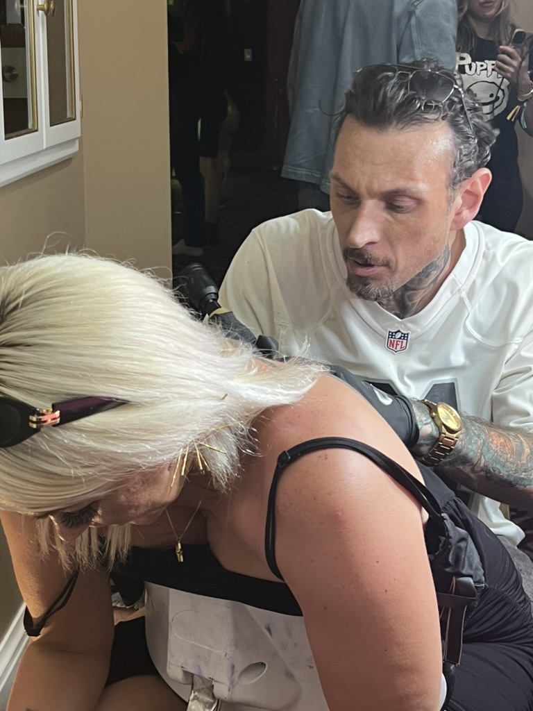 Joshua Cole tattoos upper back on blonde client leaning on portable chair at offsite — Joshua Cole offsite booking, Work of Art Las Vegas
