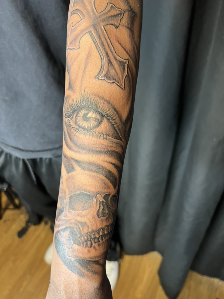 Black and grey cross, eye, and skull forearm tattoo on brown skin. — Joshua Cole, Work of Art Las Vegas