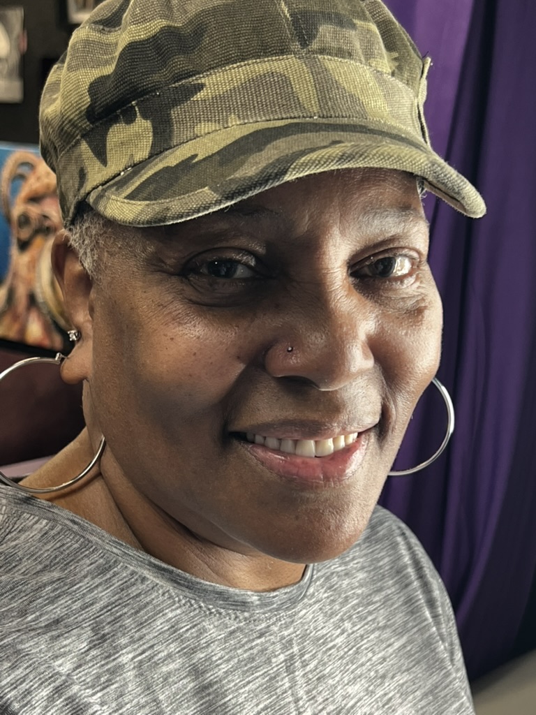 Smiling woman in camo cadet hat with small silver nostril stud and hoop earrings — Joshua Cole, Work of Art Las Vegas