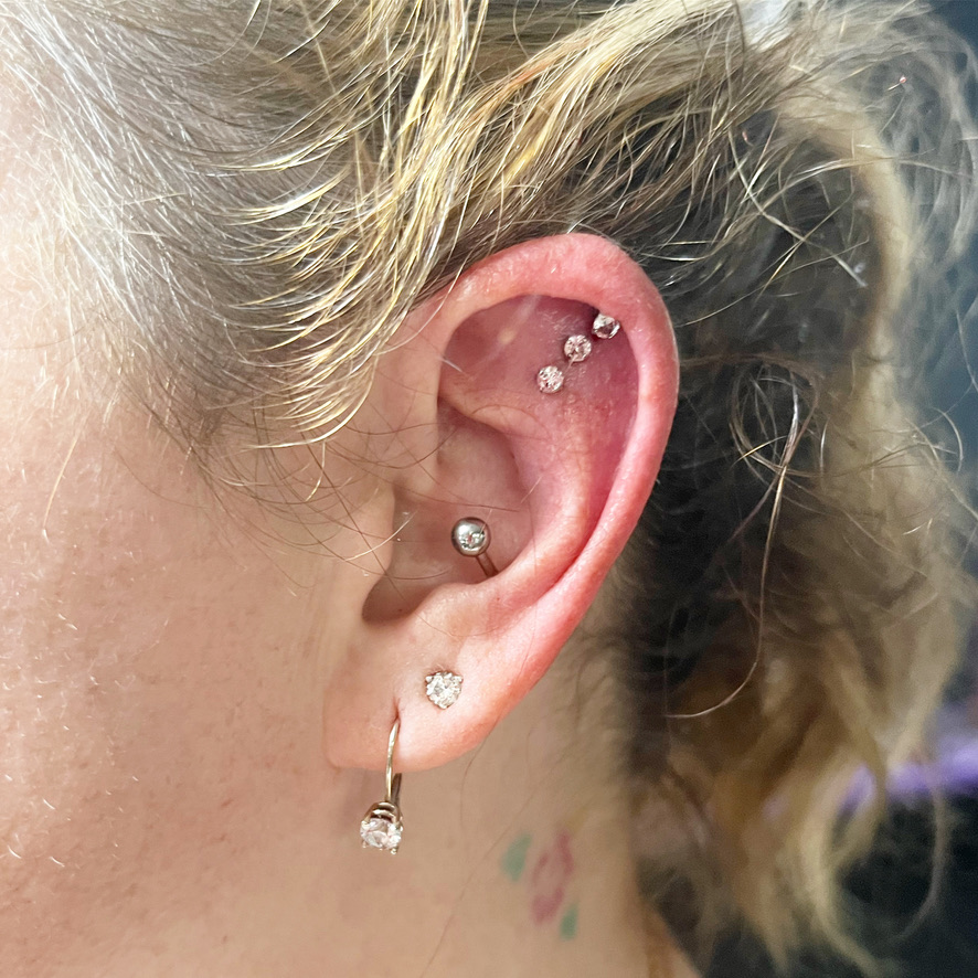 Blonde client ear with three flat helix studs, conch barbell and dangling lobe crystal — Joshua Cole, Work of Art Las Vegas